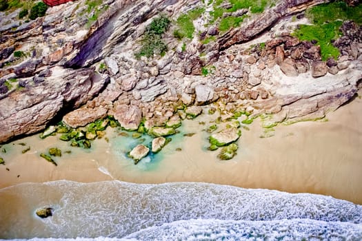 Aerial view of interesting weathered rock formations above a sandy beach with shallow surf lapping at the shore