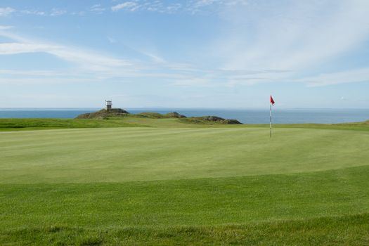 A putting green with tended grass and flag pole marker and a coast guard lookout station overlooking the sea.