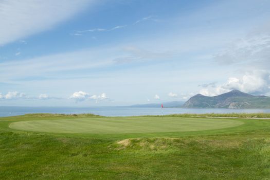 A putting green with tended grass and flag pole marker overlooking the sea and distant mountains.