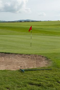 A sand bunker with rake next to a putting green with marker flag with a grey cloudy sky in the distance.