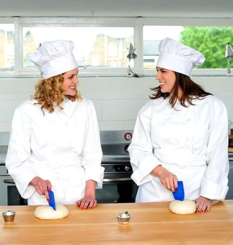 Two pretty female chefs looking at each other while kneading dough