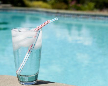 Plain glass of water with ice cubes on side of blue swimming pool
