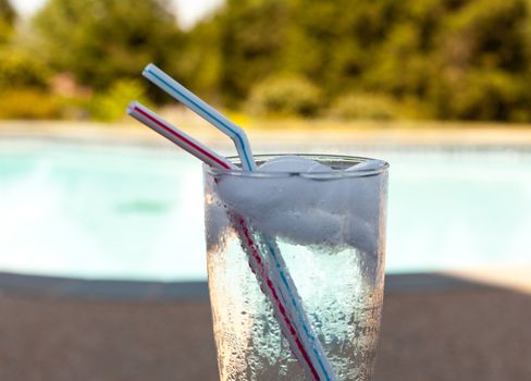 Plain glass of water with ice cubes on side of blue swimming pool