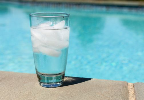 Plain glass of water with ice cubes on side of blue swimming pool