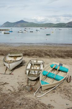 Wooden rowing boats on hand pulling trailers on a sandy beach with the sea, boats and a mountain in the distance.