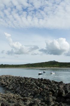 A rock breakwater shields boats moored in the water with a beach in the distance and a moody cloud sky.