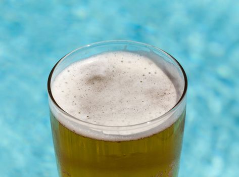 Plain pint glass of cold lager beer sitting on edge of blue swimming pool