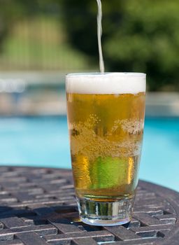 Plain pint glass of beer being poured sitting on table by blue swimming pool