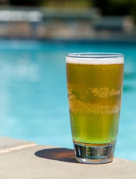 Plain pint glass of cold lager beer sitting on edge of blue swimming pool