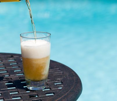 Plain pint glass of beer being poured sitting on table by blue swimming pool