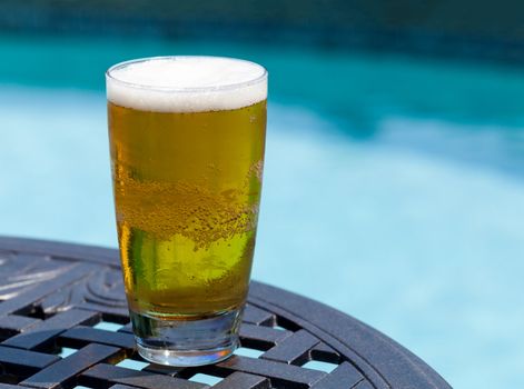 Plain pint glass of cold lager beer sitting on table by blue swimming pool