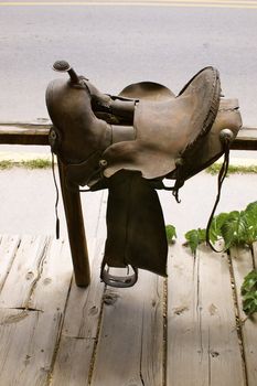 American brown leather saddle placed across a wooden bar against a rustic wooden floor with copy space.