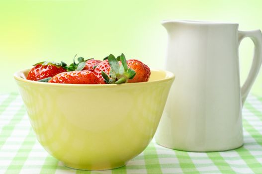 Eye level close up view of a yellow china bowl of red strawberries and a cream jug, on a green and white gingham table cloth with green, yellow and white bokeh in the background.