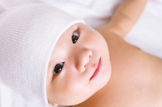 A cute little baby is looking into the camera and wearing a white hat. 