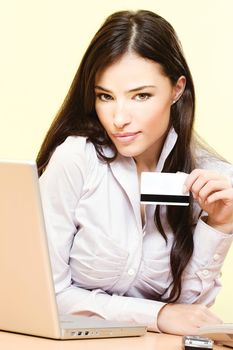 Beautiful young woman sitting near laptop and mobile phone holding credit card in her left hand