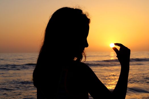 Silhouette of young girl holding the sun in her fingers at the beach during beautiful sunset