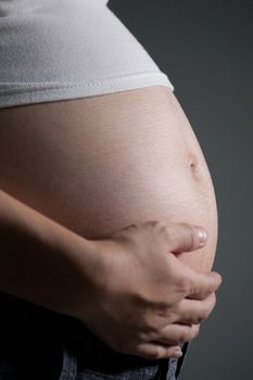 Close-up of a pregnant woman caressing her belly while standing against a gray background.