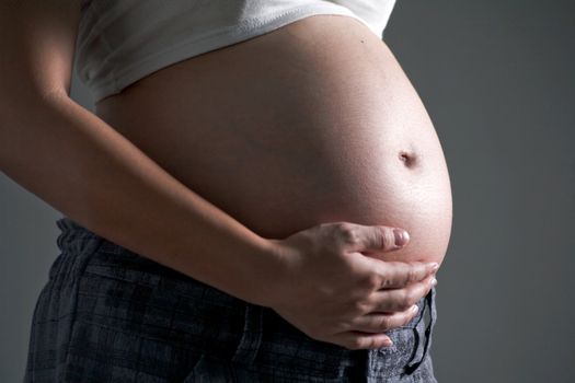 Close-up of a pregnant woman caressing her belly while standing against a gray background.