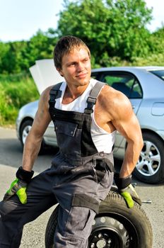 Portrait of a handsome mechanic sitting on a tire on a road.