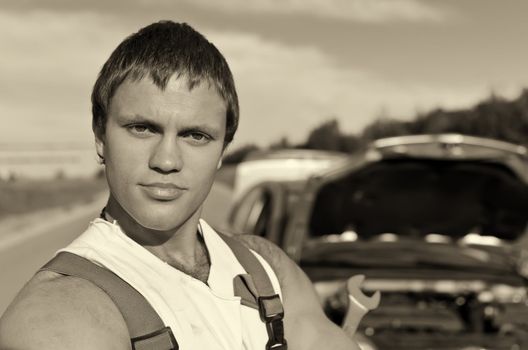 Black and white portrait of a hadsome mechanic with a broken car on background