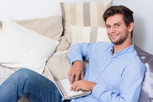 Young brunette man in blue shirt using laptop on couch