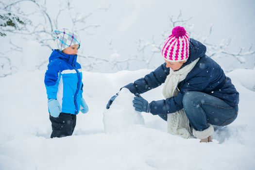 Smiling happy boy with his mother having fun outdoors on snowing winter day in Alps playing in snow.