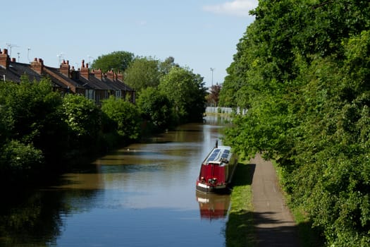 A waterway, canal, with a moored barge, boat, next to a towpath with trees and houses.