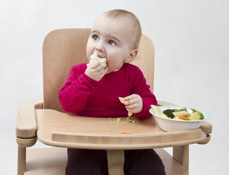 young child in red shirt eating vegetables in wooden chair.