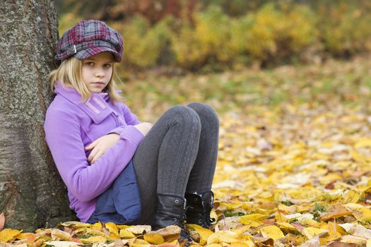 Full length portrait of a little girl sitting under tree in autumn park 