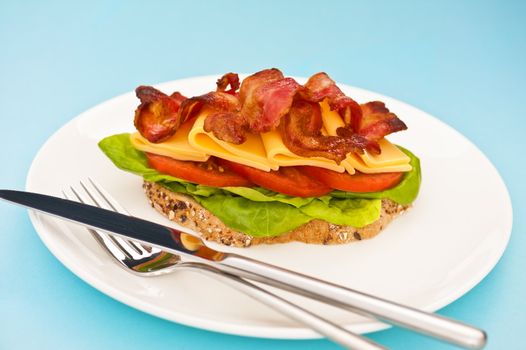 Open blt sandwich on a white plate with blue background with fork and knife