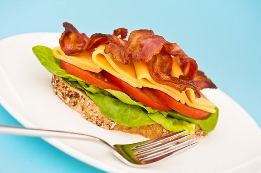 Open blt sandwich on a white plate with blue background and fork