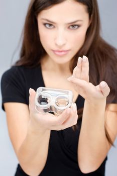 Young woman holding contact lenses cases and lens in front of her. Focus on lens and container.