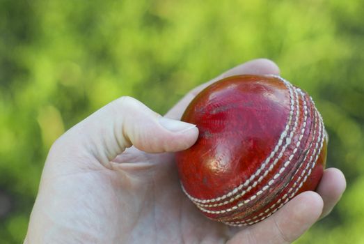 Bowler's hand holding a red leather cricket ball with green grass background