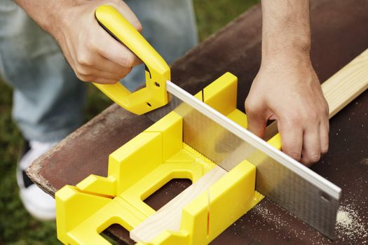 Man cutting a slat of wood using a saw and miter box outdoors.