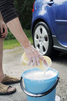 Hand lifting a sponge from a bucket. A Blue car in the background.