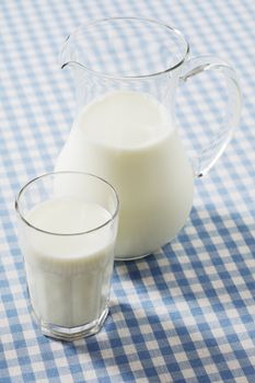 A glass of milk and a milk jug on plaid tablecloth.