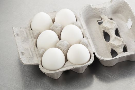 Six white hen eggs in a carton on a scratched metal surface.