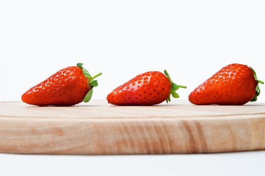 A line of three red strawberries with leaves and short stalks intact, shot sideways at eye level, on a wooden chopping board against an off-white background.