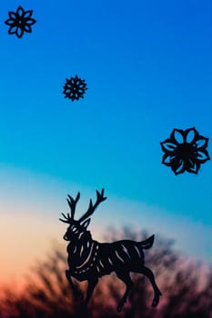 Silhouette of a deer in wood under falling snowflakes. Application from a paper on window glass