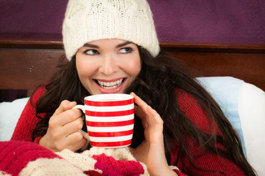 A beautiful smiling young woman enjoying a hot frink under the blankets in bed.