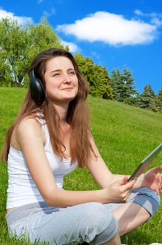 Smiling happy girl with headphones and tablet sitting on grass in park.