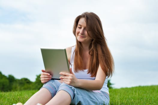 Happy girl with tablet sitting on grass in park.