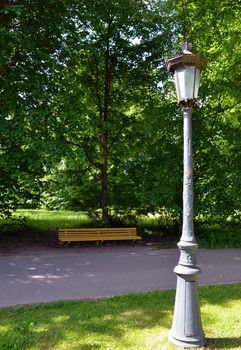 Vintage retro park lamp light pole and yellow wooden bench under trees for recreation.