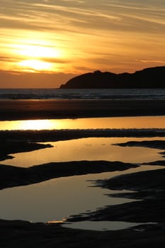 pools of water on a beach leads to the sea with a silhouette headland with a cloud sunset in the distance.