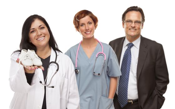 Attractive Hispanic Female Doctor or Nurse Holding Out Baby Shoes and Support Staff Behind Isolated on a White Background.