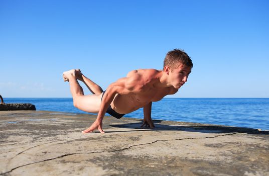 Young strong teenage athlete doing exercise on pier near sea under blue sky