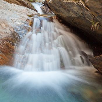Small natural waterfall casdcading over rocks into a calm blue pond below with silky appearance from long exposure
