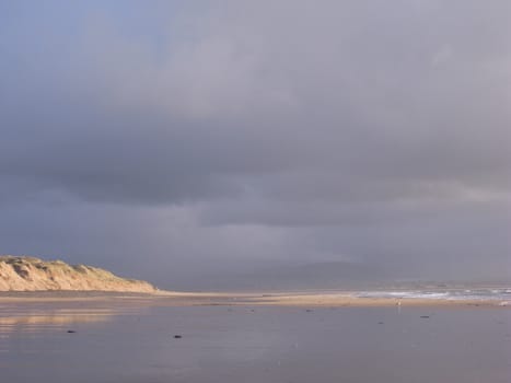 A beach with the incoming tide and a sand dune lit up by the sun with a storm cloud in the distance.