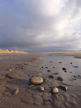 A beach with pebbles in the foreground amongst rippled sand and a dune and storm cloud in the distance.