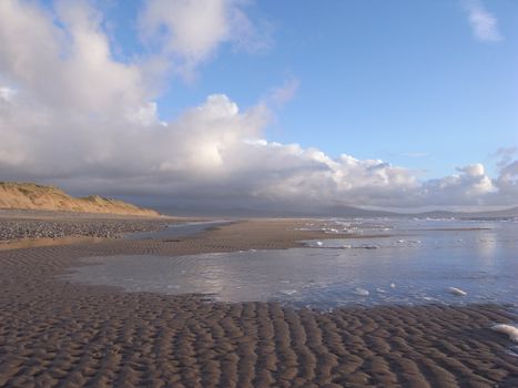 A sandy beach with rippled sand and a dune with the tide bringing the sea over the sand with reflections of a blue cloudy sky.